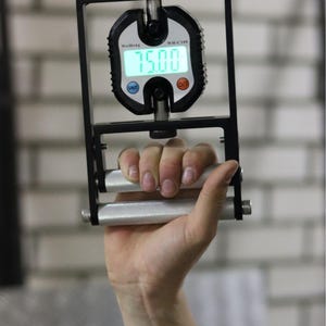 Può includere: Un dinamometro per impugnatura nero e argento con display digitale che mostra "75.00". Il dispositivo è tenuto in una mano, con il display e le maniglie visibili. È progettato per misurare la forza di presa.