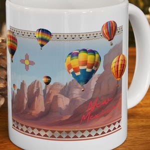 May include: A white ceramic mug featuring a colorful graphic of hot air balloons floating over a desert landscape with rock formations. The design includes the text New Mexico and a Zia sun symbol, framed by decorative geometric patterns.