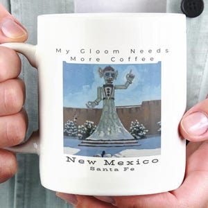 May include: A white ceramic mug featuring a graphic of a statue in a snowy landscape with the text My Gloom Needs More Coffee above the image and New Mexico Santa Fe printed below the image in black lettering on the front of the mug.