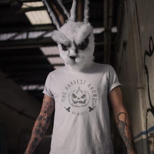 May include: A person wearing a white rabbit mask and a light gray t-shirt. The shirt features a pumpkin graphic with the text "THE HARVEST KNOWS NO MERCY". The person has tattoos on their arms.