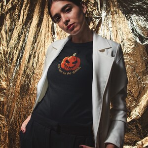 May include: A black t-shirt with a jack-o'-lantern graphic and the text "just here for the gourds." The person is wearing a white blazer and black pants. The background is a textured gold surface.