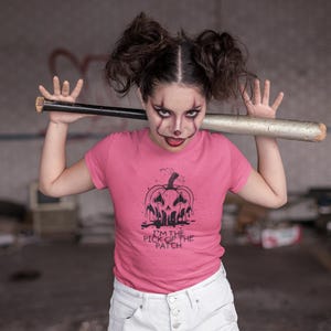 May include: A pink t-shirt with a black pumpkin graphic and the text "I'M THE PICK OF THE PATCH." A baseball bat rests on the shoulders. The person is wearing white shorts and has clown-like makeup.
