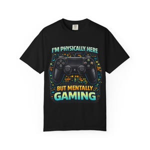 Puede incluir: Camiseta negra con un diseño de un mando de videojuegos negro. El texto en la camiseta dice "I'M PHYSICALLY HERE BUT MENTALLY GAMING" en blanco y turquesa. El diseño tiene un fondo pixelado en naranja, turquesa y amarillo.