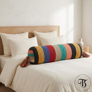Puede incluir: Un cojín cilíndrico y colorido con un patrón de rayas en varios colores, incluyendo negro, amarillo, azul, rojo y rosa. El cojín tiene borlas en cada extremo y está colocado sobre una cama con ropa de cama blanca.