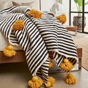 Puede incluir: Una manta a rayas blancas y negras con borlas y pompones amarillos. La manta está colocada sobre una cama, mostrando su textura gruesa y suave. Las borlas y los pompones añaden un elemento decorativo al acogedor artículo de decoración del hogar.