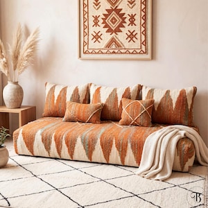 May include: A floor sofa with orange and cream geometric patterned upholstery and matching throw pillows. A cream-colored blanket is draped over the sofa. A framed wall hanging with a similar design hangs above the sofa. A cream and black patterned rug is in the foreground.