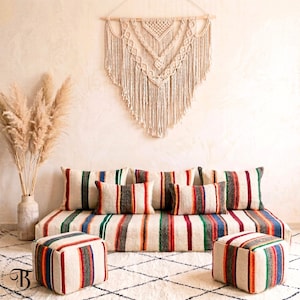 Peut inclure: Un salon de style bohème avec un canapé rayé, des coussins assortis et deux poufs carrés. Une tenture murale en macramé et un vase d'herbe de la pampa séchée complètent le décor. La palette de couleurs comprend crème, rouge, vert et bleu.