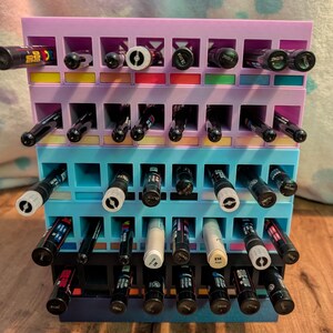 Puede incluir: Un organizador de bolígrafos y rotuladores de varios niveles en tonos morado, azul y negro. El organizador está lleno de varios bolígrafos y rotuladores, mostrando una gama de colores y estilos de punta. El diseño incluye compartimentos individuales.