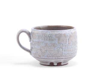 May include: A ceramic mug with a light blue and white floral design. The mug has a handle and a textured surface with embossed floral patterns. The rim and base have a darker, brownish colour.