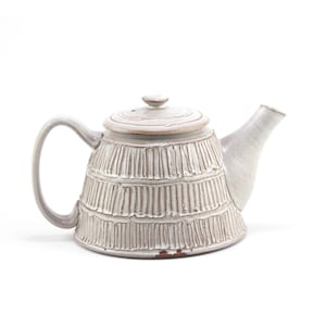 May include: A ceramic teapot with a light gray glaze and a textured surface featuring vertical ridges arranged in horizontal bands. The teapot includes a matching lid with a small knob, a curved handle, and a tapered spout.