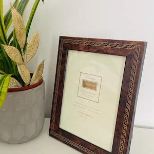 May include: A rectangular picture frame with a dark brown burl wood finish and a decorative geometric inlay border. The frame contains a product insert labeled Debenhams Fine Frames, noting dimensions of 5x7 inches with mount and 6x8 inches without.