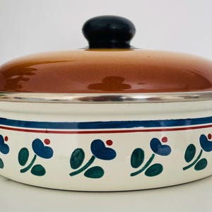 May include: A vintage casserole dish with a brown lid and black knob. The off-white body features a blue and green floral pattern with a blue and red stripe. It has wooden handles.