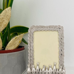 May include: A rectangular silver-toned picture frame featuring a braided border design. The bottom edge of the frame is decorated with a row of ten small, stylised cat figures of varying heights standing side-by-side in a line.