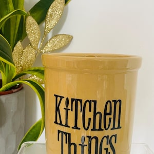 May include: A tan ceramic kitchen utensil holder with the words "Kitchen Things" in black lettering. The holder is cylindrical and sits on a clear surface. A potted plant and gold glittery leaves are in the background.