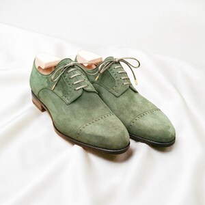 May include: A pair of green suede dress shoes with a classic Oxford design. The shoes feature a cap toe, detailed stitching, and matching green laces. Wooden shoe trees are inserted inside the shoes. The shoes are displayed on a white background.