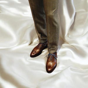 May include: A pair of brown leather dress shoes with blue laces, worn with brown trousers. The shoes feature a cap-toe design. The image is set against a white fabric background, creating a classic and elegant aesthetic.