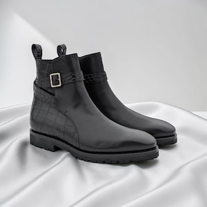 May include: A pair of black leather ankle boots with a side zip and buckle fastening. The boots feature a textured, crocodile-effect panel and a pull tab. The soles are black with a lugged design. The boots are displayed on a white satin surface.