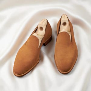 Pode incluir: Um par de mocassins de camurça castanhos com formas de madeira no interior. Os sapatos têm um design clássico de enfiar, com bico redondo e salto baixo. São exibidos numa superfície de cetim branco.