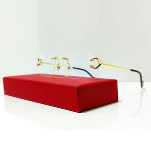 May include: Rimless spectacles with gold-coloured frames and temples, resting on a red rectangular box. The spectacles have a clear lens and a distinctive design. The background is plain white.