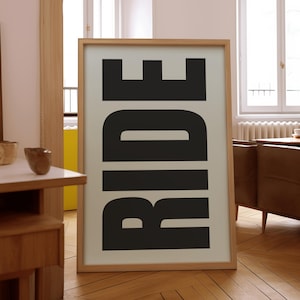 Könnte beinhalten: Ein gerahmter Kunstdruck mit dem Wort "RIDE" in großen, fetten, schwarzen Buchstaben auf cremefarbenem Hintergrund. Der Rahmen ist aus hellem Holz. Der Druck wird auf einem Holzboden präsentiert.