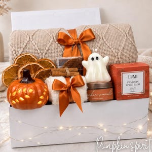 May include: A white gift box containing a beige textured throw blanket tied with an orange ribbon, dried orange slices, a ceramic orange pumpkin, a white ghost-shaped candle, a small candle tin, and a box of Lumi Pumpkin Spice Chai tea.