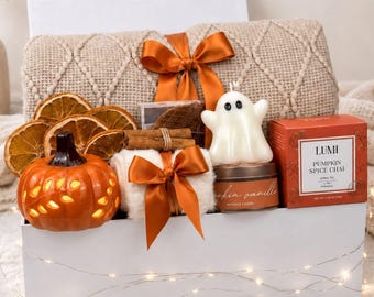 Op de afbeelding: Een witte geschenkdoos met een beige, getextureerde deken vastgebonden met een oranje lint, gedroogde sinaasappelschijfjes, een oranje keramische pompoen, een witte kaars in de vorm van een spookje, een kaarsblikje en Lumi Pumpkin Spice Chai thee.