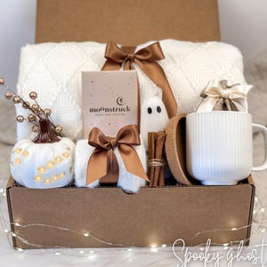 May include: A gift box containing a white textured blanket with a brown ribbon, a Moonstruck Portland Oregon box, a white ceramic pumpkin with cutouts, a small ghost figurine, cinnamon sticks, a cork coaster, and a white ribbed mug with a bow.