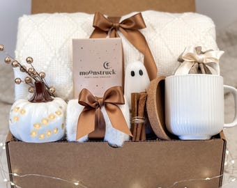Op de afbeelding: Een geschenkdoos met een witte, getextureerde deken met een bruin lint, een Moonstruck Portland Oregon doos, een witte keramische pompoen met uitsparingen, een klein spookfiguurtje, kaneelstokjes, een kurken onderzetter en een witte geribbelde mok.