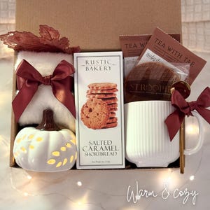 May include: A gift box filled with a white pumpkin-shaped candle holder, a white mug, cookies, tea bags, and a fluffy white item tied with a burgundy bow. The box is decorated with burgundy accents and the words "Warm & cozy".