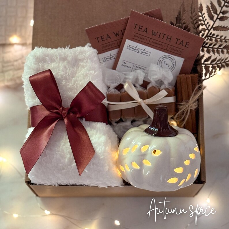 May include: A gift box with a white plush towel tied with a burgundy ribbon, tea packets, wrapped candies, cinnamon sticks, and a white pumpkin-shaped candle holder. The box is labeled "Autumn spice".