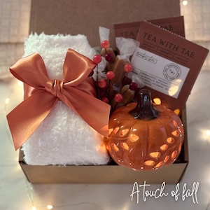 May include: A gift box with a white fluffy item tied with an orange satin bow, a ceramic pumpkin, tea packets, and decorative berries. The box is labeled "A touch of fall".