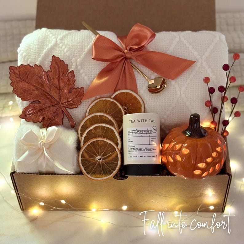 May include: A gift box with a fall theme, featuring a white blanket, orange bow, decorative leaf, dried orange slices, a candle, and a pumpkin-shaped ceramic holder. The box is decorated with fairy lights and the text "Fall into comfort".