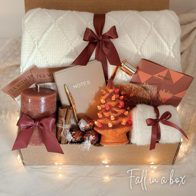 May include: A gift box containing a white diamond-patterned throw blanket tied with a brown ribbon, a tan notebook labeled NOTES, a brown pillar candle with a brown ribbon, cinnamon sticks, chocolates, a ceramic tree with lights, and fuzzy socks.