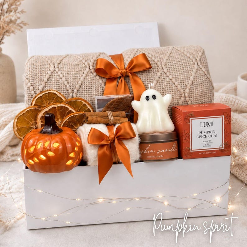 Op de afbeelding: Een witte geschenkdoos met een beige, getextureerde deken vastgebonden met een oranje lint, gedroogde sinaasappelschijfjes, een oranje keramische pompoen, een witte kaars in de vorm van een spookje, een kaarsblikje en Lumi Pumpkin Spice Chai thee.