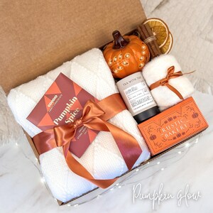 May include: A gift box filled with fall-themed items. Includes a white blanket, a pumpkin-shaped candle, a box of chocolate, a tea tube, a small white towel, and a box of cookies. The box is tied with a rust-colored ribbon. The text "Pumpkin Spice" is visible.