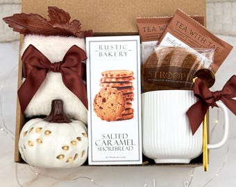 May include: A gift box containing a white fuzzy item with a brown ribbon, a white ceramic pumpkin, a box of Rustic Bakery Salted Caramel Shortbread, two Tea With Tae packets, a Stroopwafel, and a white ribbed mug with a gold spoon and brown ribbon.