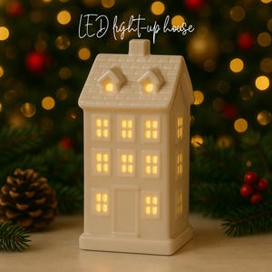 May include: A cream-colored LED light-up house with illuminated windows and the text "LED light-up house". The house has a pitched roof and a chimney. The background features blurred Christmas lights and greenery, creating a festive atmosphere.
