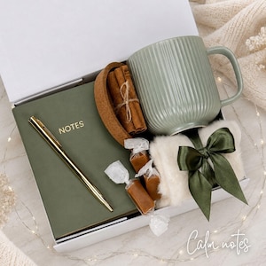 Op de afbeelding: Een geschenkdoos met een saliegroene geribbelde keramische mok, een donker olijfgroen notitieboek met het woord NOTES in goud, een gouden pen, een kurken onderzetter met kaneelstokjes, twee verpakte karamelsnoepjes en een wit imitatiebont met een groene strik.