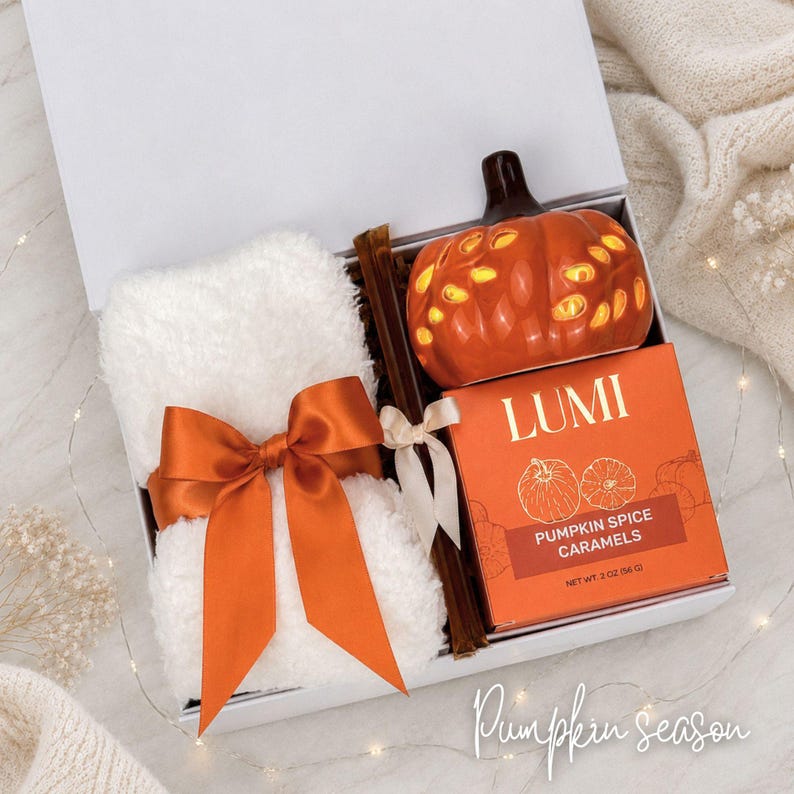 May include: A white gift box containing a folded white plush blanket tied with an orange satin ribbon, a brown glass stirrer, a ceramic orange pumpkin-shaped light, and a box of Lumi Pumpkin Spice Caramels with a net weight of 2 ounces.