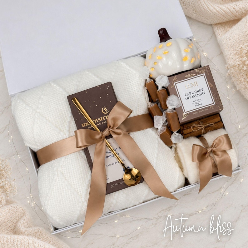 May include: A gift box containing a white cable-knit blanket, a dark chocolate bar with a gold spoon, a white ceramic pumpkin, a box of Earl Grey Moonlight tea, cinnamon sticks, and small wrapped candies, all tied with a brown satin ribbon.