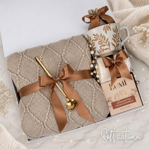 May include: A gift box containing a beige knit blanket with a diamond pattern, a gold-toned spoon, a ceramic mug with a gold leaf design, and a box of Lumi handcrafted pecan caramels, all tied with brown satin ribbons and accented with pearls.