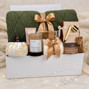 Op de afbeelding: Een geschenkset in een witte doos met een donkergroene, getextureerde gebreide deken met een gouden lint, een witte keramische pompoen, een Tea With Tae-koker, een Moonstruck-doos met een gouden hulpmiddel, een kaneelsuikerkaars en kaneelstokjes.