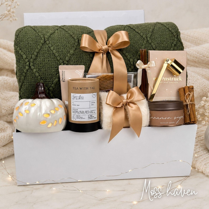Op de afbeelding: Een geschenkset in een witte doos met een donkergroene, getextureerde gebreide deken met een gouden lint, een witte keramische pompoen, een Tea With Tae-koker, een Moonstruck-doos met een gouden hulpmiddel, een kaneelsuikerkaars en kaneelstokjes.