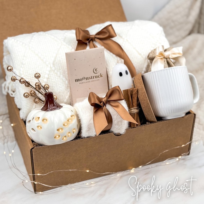 May include: A gift box filled with a white cable-knit blanket, a ceramic pumpkin, a white mug, a small ghost figurine, and a book. The box is tied with brown satin ribbons. The text "Spooky ghost" is at the bottom.