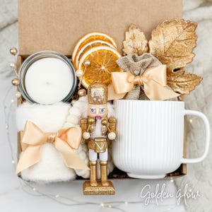 May include: A gift box with a white mug, candle, and a gold-colored nutcracker. The box also contains dried orange slices, a small burlap bag with a bow, and a white blanket with a satin bow. The text "Golden glow" is at the bottom right.