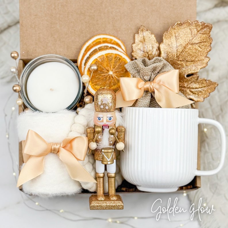 May include: A gift box with a white mug, candle, and a gold-colored nutcracker. The box also contains dried orange slices, a small burlap bag with a bow, and a white blanket with a satin bow. The text "Golden glow" is at the bottom right.