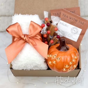 May include: A gift box with fall-themed items. It includes a white fluffy item tied with an orange bow, a ceramic pumpkin with cut-out designs, tea packets, and caramel candies. The box is decorated with a sprig of red berries and the text "A touch of fall".