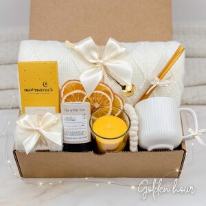 May include: A gift box with a white blanket, mug, candle, tea, and dried orange slices. The box is tied with a cream-colored bow. The box contains a yellow candle and a white mug with vertical lines.
