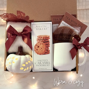 May include: A gift box containing a white fuzzy item with a burgundy ribbon, a white ceramic pumpkin, a box of Rustic Bakery Salted Caramel Shortbread, a Stroopwafel, Tea With Tae tea bags, and a white ribbed mug with a gold spoon and burgundy ribbon.