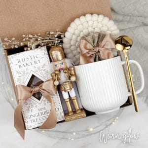 May include: A gift box with a white mug, gold spoon, and a miniature nutcracker. The box also contains a package of mini gingerbread cookies, a beige bow, and a decorative coaster. The words "Warm glow" are written in cursive on the bottom right.